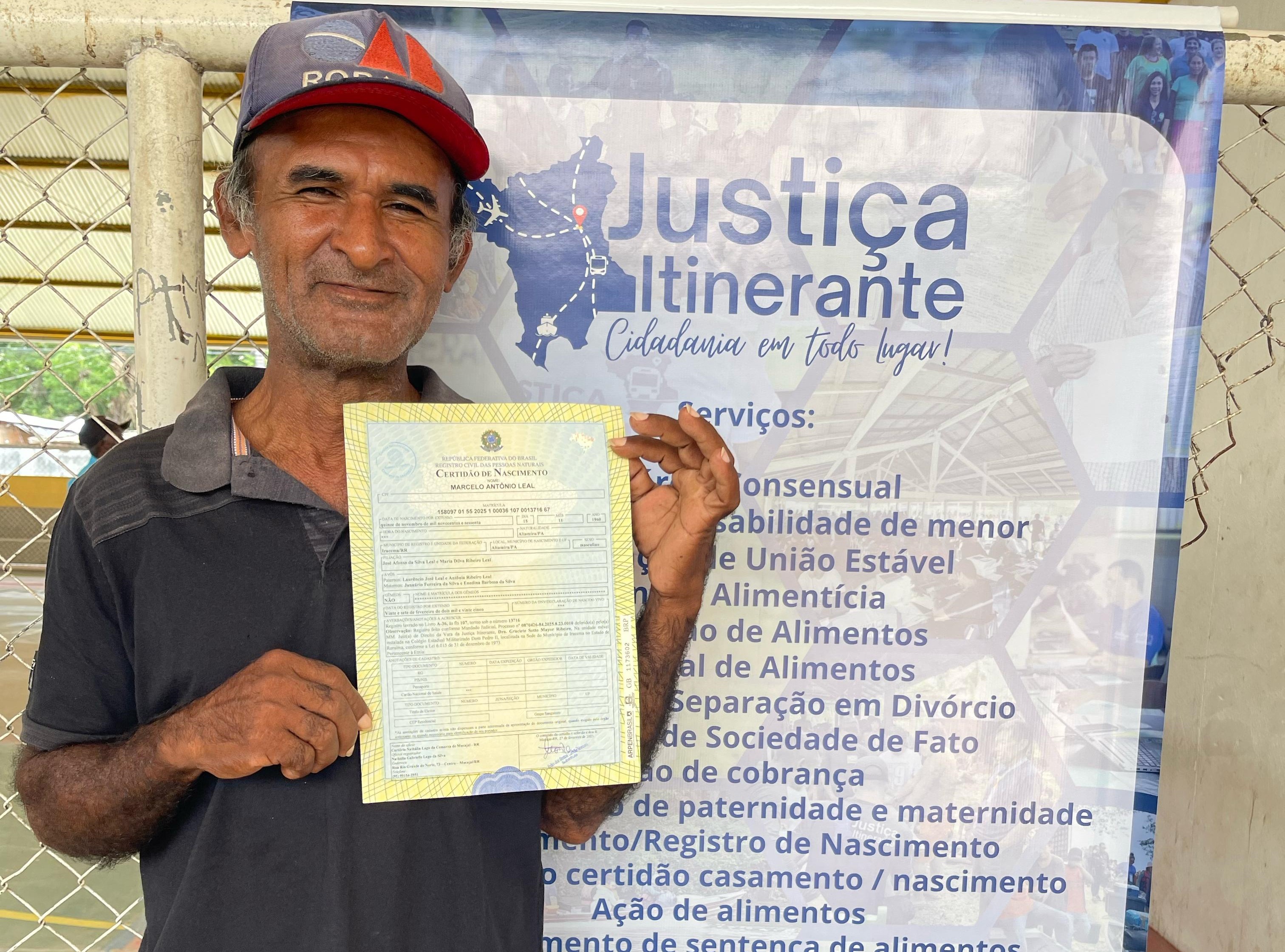 Imagem colorida contém o cidadão, Leandro Leal, atendido pela Vara da Justiça Itinerante do TJRR, segurando um certificado de nascimento. Atrás dele encontra-se um banner da Justiça Itinerante contendo os serviços da Justiça Itinerante listados.