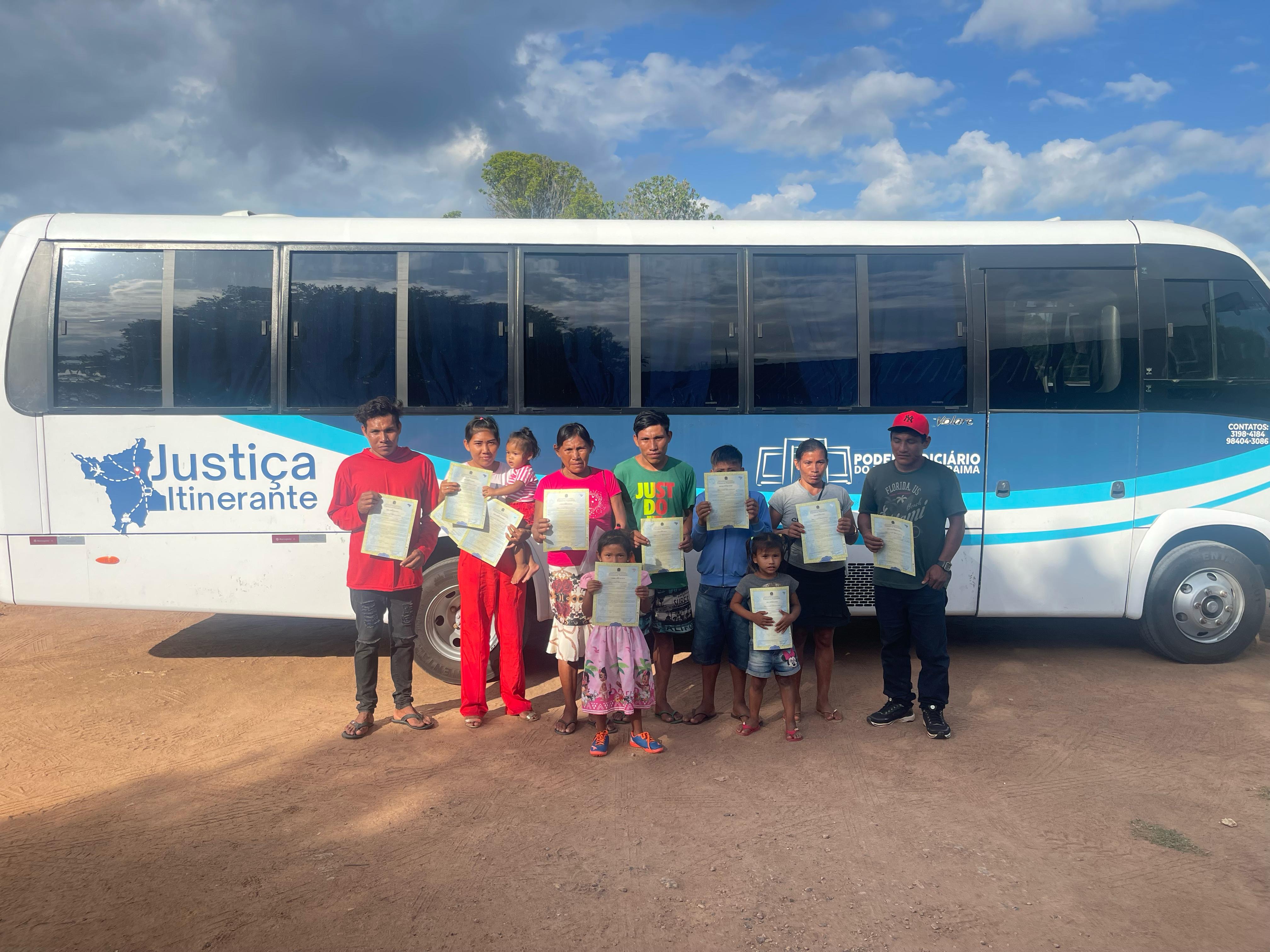 Foto colorida mostrando 10 pessoas, entre crianças, adultos e idosos, em frente ao onibus da Justiça Itinerante, segurando as certidões de nascimento
