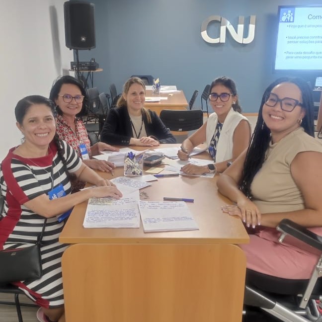 Imagem colorida mostra participantes em um evento no Conselho Nacional de Justiça (CNJ), reunidos em uma mesa de trabalho com anotações e materiais de estudo. O grupo é composto por mulheres que interagem, escrevem e sorriem para a câmera, entre elas a Desembargadora Tânia Vasconcellos e a servidora Claudete Pereira. Ao fundo, é possível ver o logotipo do CNJ na parede e uma tela com uma apresentação.