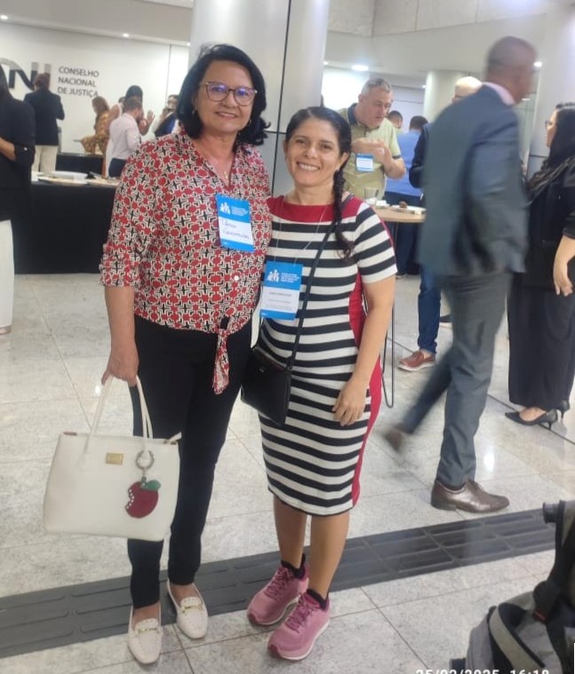 Imagem colorida mostra a Desembargadora Tânia Vasconcellos e a servidora Claudete Pereira,participantes do evento posando para uma foto no saguão do Conselho Nacional de Justiça (CNJ), Ambas utilizam crachás de identificação e sorriem para a câmera. No ambiente, há outras pessoas circulando e interagindo. Ao fundo, o nome do Conselho Nacional de Justiça está visível na parede.
