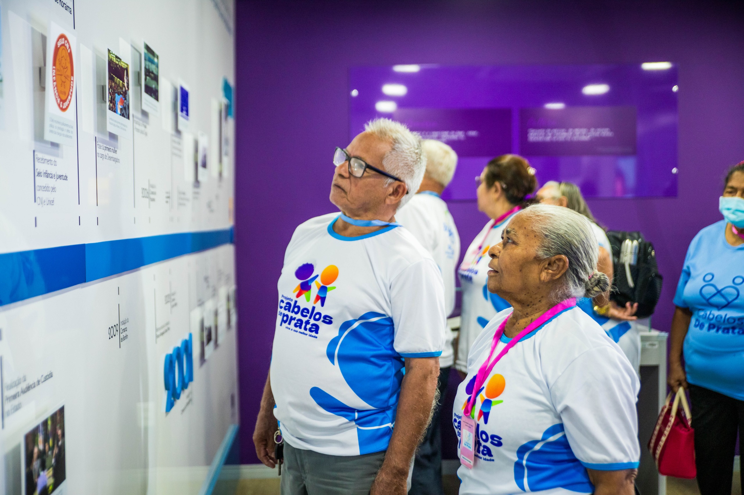 imagem colorida contém um casal de idosos observando a linha do tempo destacada na parede do CMC/TJRR, durante visita do grupo cabelos de prata.