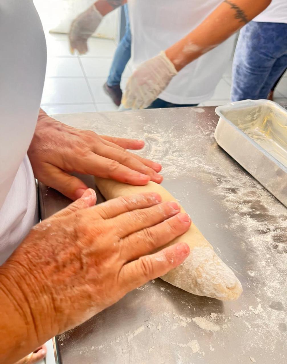 foto colorida em plano fechado dando enfoque em uma mão amassando a massa do pão. Abaixo está o texto “Projetos Sociais da VEPEMA: Natudivas e Impactos previnem violência e auxiliam mulheres”.