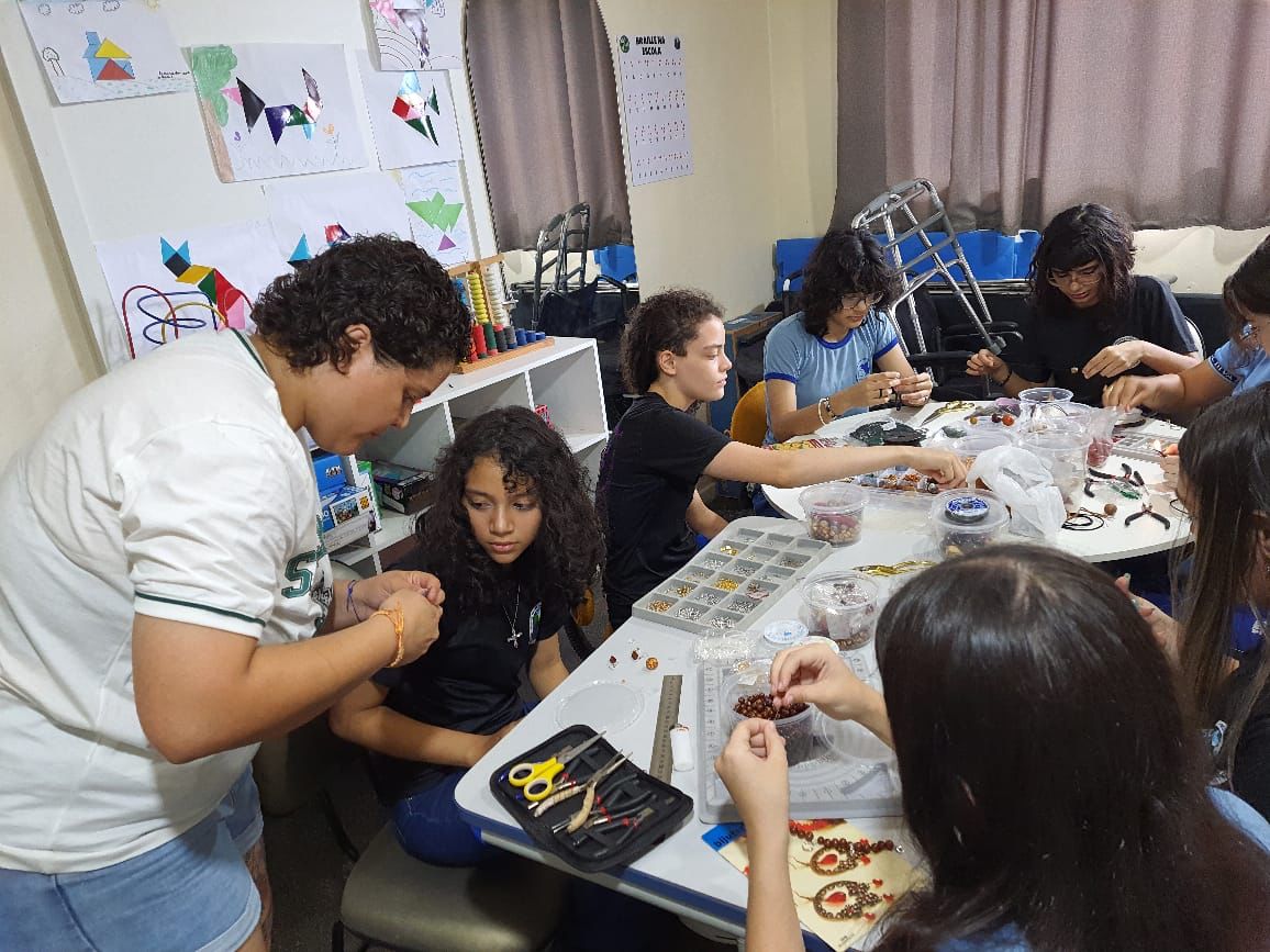 foto colorida de um grupo de alunas mulheres sentadas ao redor de uma mesa, produzindo as biojoias. Abaixo está o texto “Projetos Sociais da VEPEMA: Natudivas e Impactos previnem violência e auxiliam mulheres”