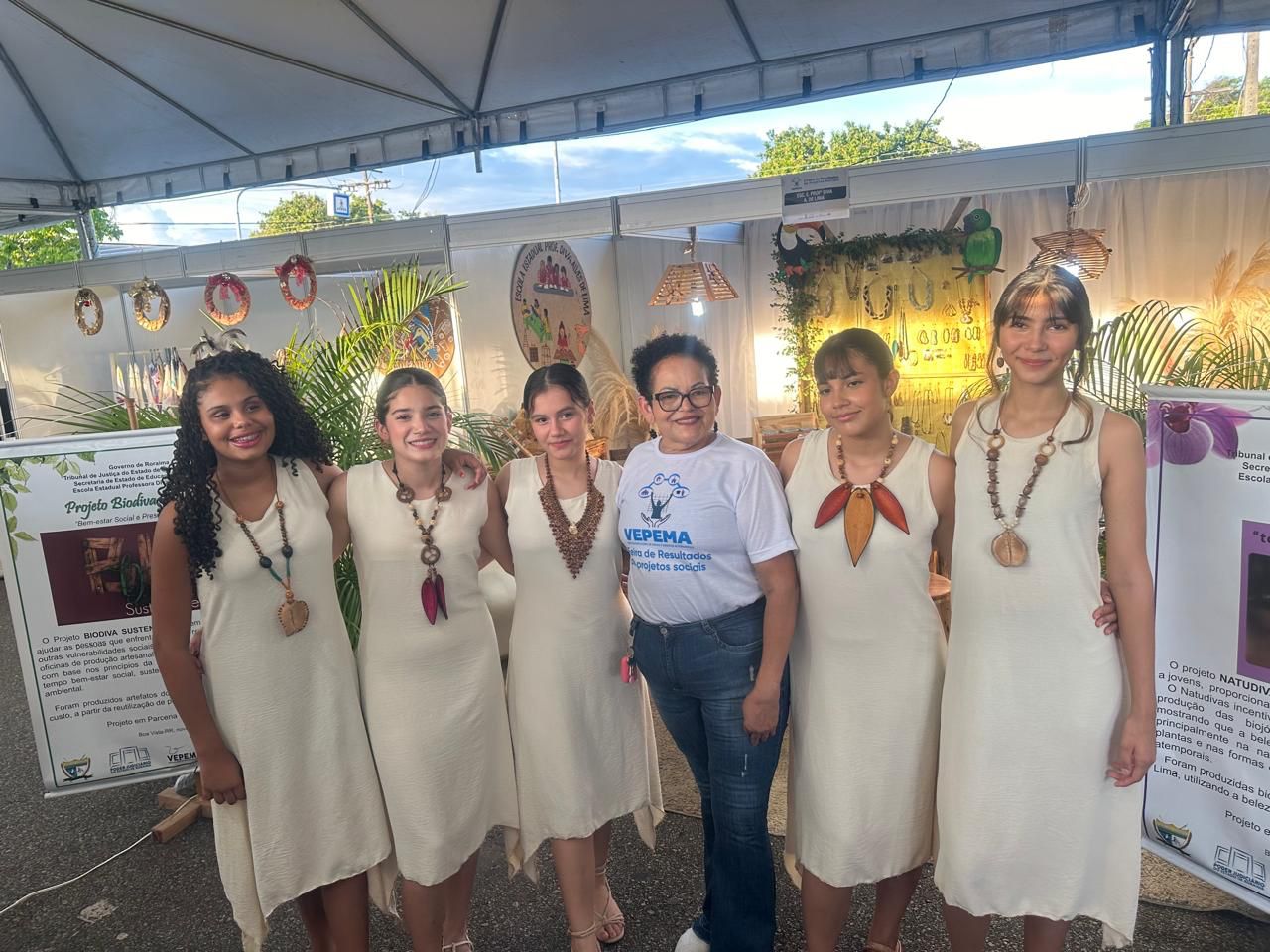 foto colorida de cinco alunas e a gestora da Escola Estadual Diva Lima posando para fotografia durante a feira de resultados da Vepema. Elas estão usando os colares produzidos. Abaixo está o texto “Projetos Sociais da VEPEMA: Natudivas e Impactos previnem violência e auxiliam mulheres”.