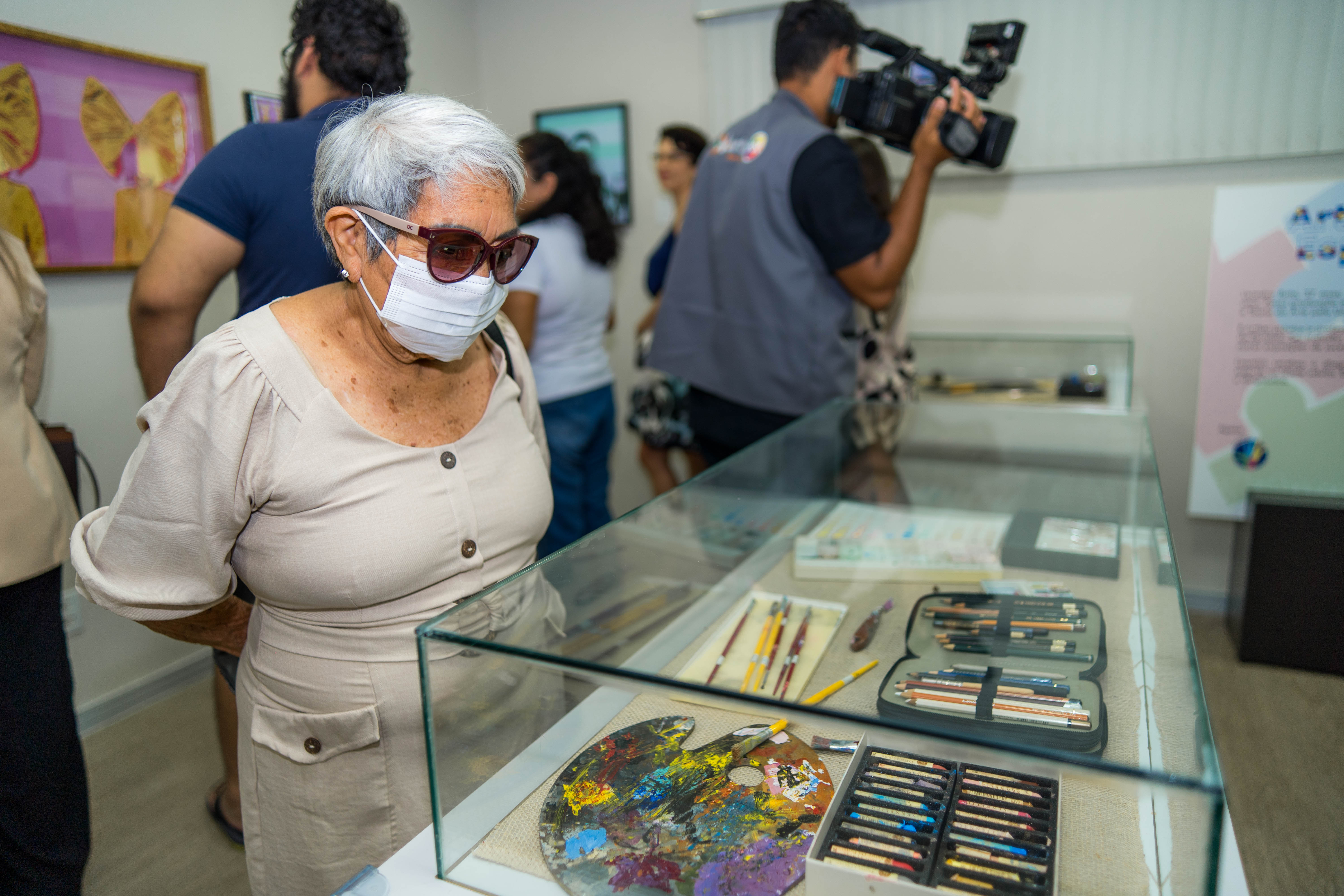 A imagem mostra o interior de uma sala de exposição, onde um grupo de pessoas observa itens em uma vitrine de vidro. Uma senhora com cabelos brancos curtos, óculos escuros e uma máscara facial branca está em primeiro plano, olhando atentamente para o conteúdo da vitrine. Dentro da vitrine, há diversos materiais de arte, incluindo uma paleta de pintura colorida com tinta seca, pincéis de diferentes tamanhos, estojos com lápis de cor e outros materiais de desenho. A imagem mostra o interior de uma sala de exposição, onde um grupo de pessoas observa itens em uma vitrine de vidro. Uma senhora com cabelos brancos curtos, óculos escuros e uma máscara facial branca está em primeiro plano, olhando atentamente para o conteúdo da vitrine. Dentro da vitrine, há diversos materiais de arte, incluindo uma paleta de pintura colorida com tinta seca, pincéis de diferentes tamanhos, estojos com lápis de cor e outros materiais de desenho.