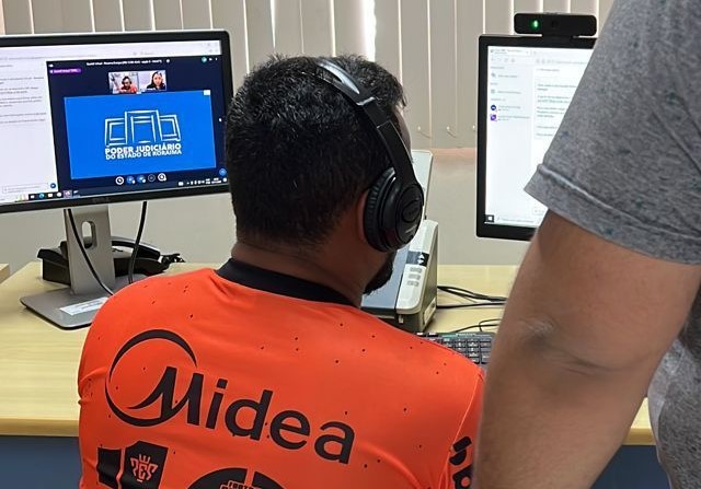 A imagem mostra um homem de costas, usando um fone de ouvido preto, sentado em frente a dois monitores de computador em um ambiente de escritório. Ele veste uma camisa laranja. A imagem mostra um homem de costas, usando um fone de ouvido preto, sentado em frente a dois monitores de computador em um ambiente de escritório. Ele veste uma camisa laranja.