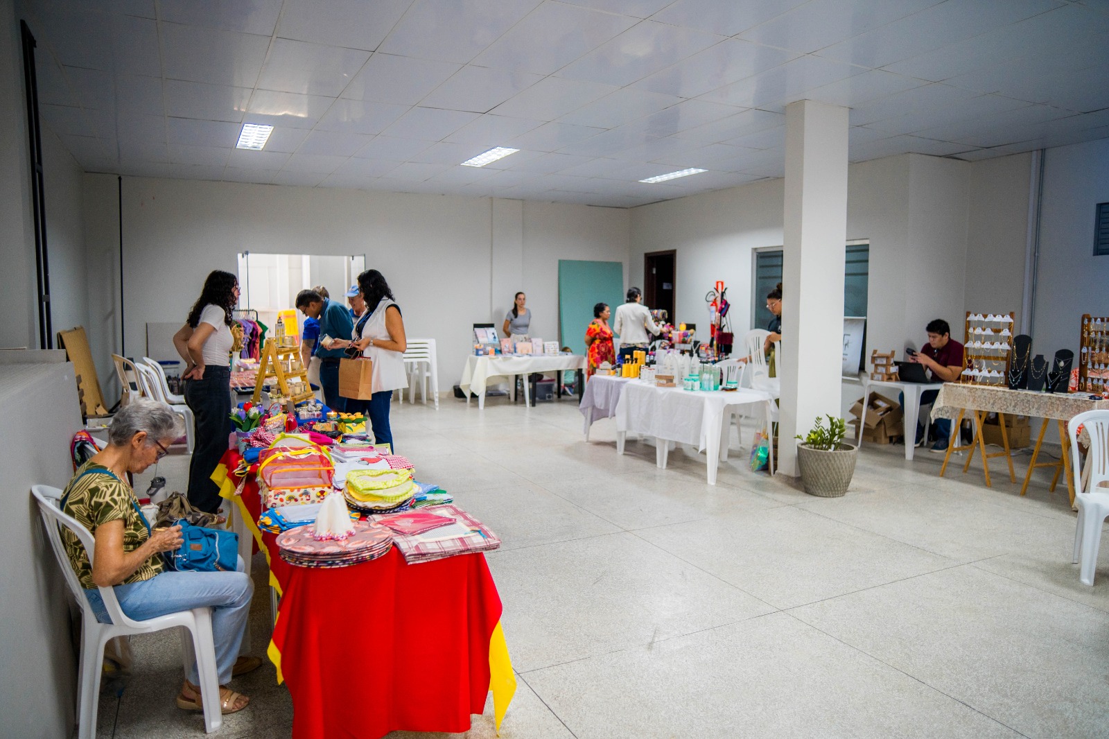  Imagem colorida contém a imagem de uma feirinha de artesanato do TJRR que ocorreu no subsolo do Palácio da Justiça. Na imagem é possivel ver diversas pessoas comprando nas exposições e vendedores sentados ou interagindo com público.
