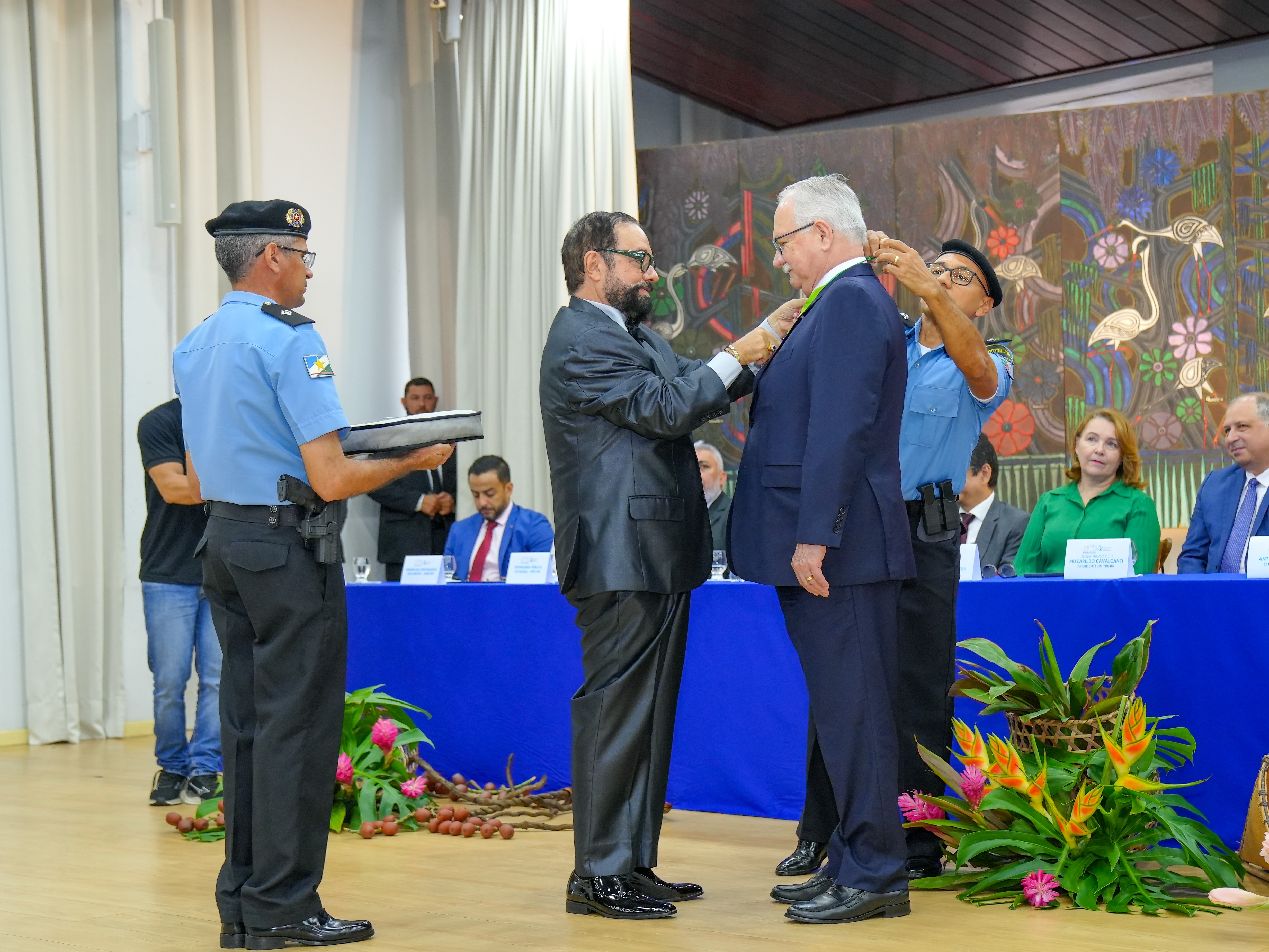 Foto colorida mostra o presidente do TJRR, desembargador Leonardo Cupello, entregando a Medalha do Mérito Judiciário ao ministro do STF, Edson Fachin, com o auxílio de dois policiais militares. Ao fundo, vê-se a mesa de autoridades. Foto colorida mostra o presidente do TJRR, desembargador Leonardo Cupello, entregando a Medalha do Mérito Judiciário ao ministro do STF, Edson Fachin, com o auxílio de dois policiais militares. Ao fundo, vê-se a mesa de autoridades.