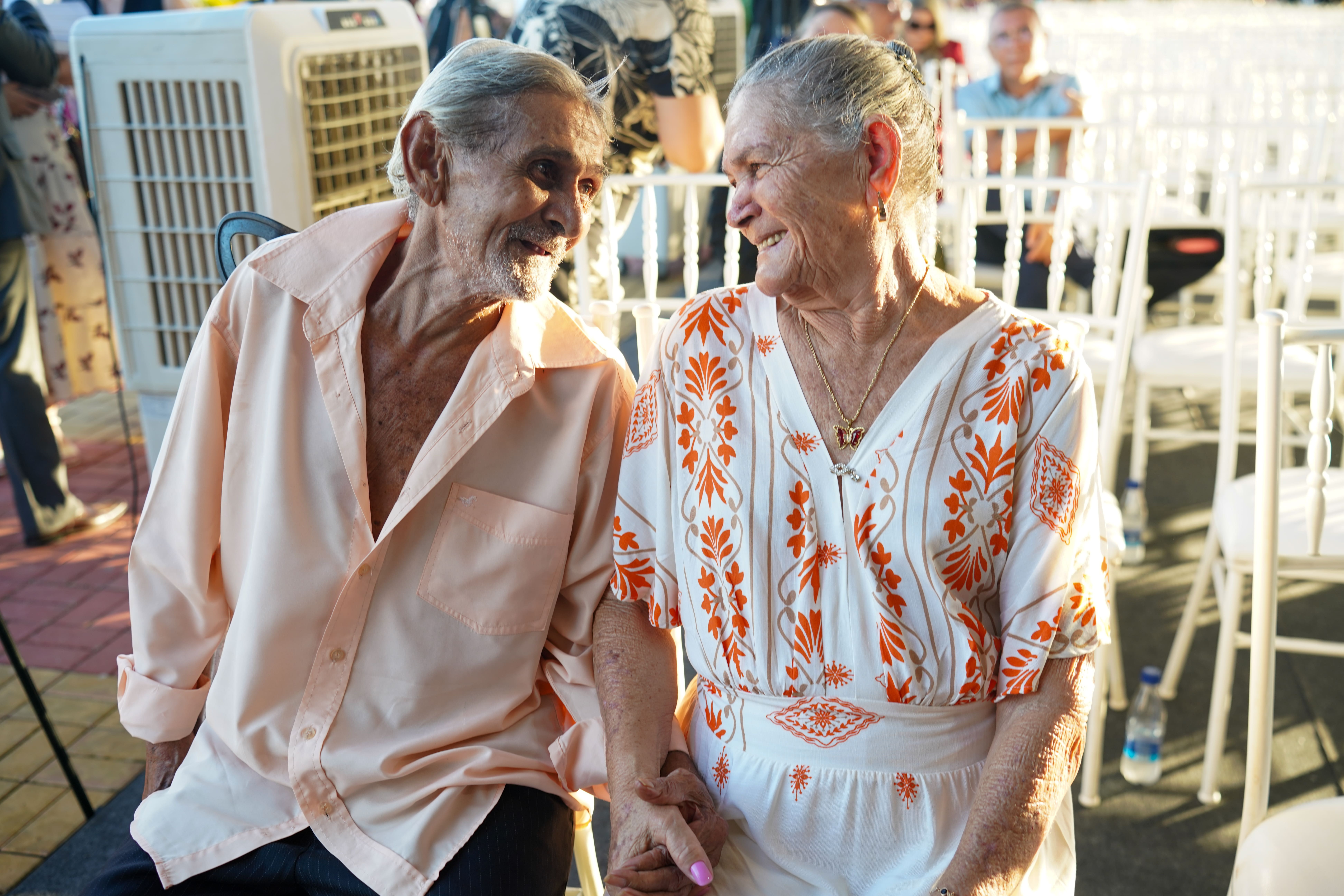 Foto colorida mostra um casal idoso sorrindo um para o outro e de mãos dadas. O homem, à esquerda, usa uma camisa social cor de pêssego. A mulher, à direita, veste uma blusa branca com um padrão floral laranja. Eles estão sentados ao ar livre, com cadeiras brancas. O sol da tarde ilumina a cena. Foto colorida mostra um casal idoso sorrindo um para o outro e de mãos dadas. O homem, à esquerda, usa uma camisa social cor de pêssego. A mulher, à direita, veste uma blusa branca com um padrão floral laranja. Eles estão sentados ao ar livre, com cadeiras brancas. O sol da tarde ilumina a cena.