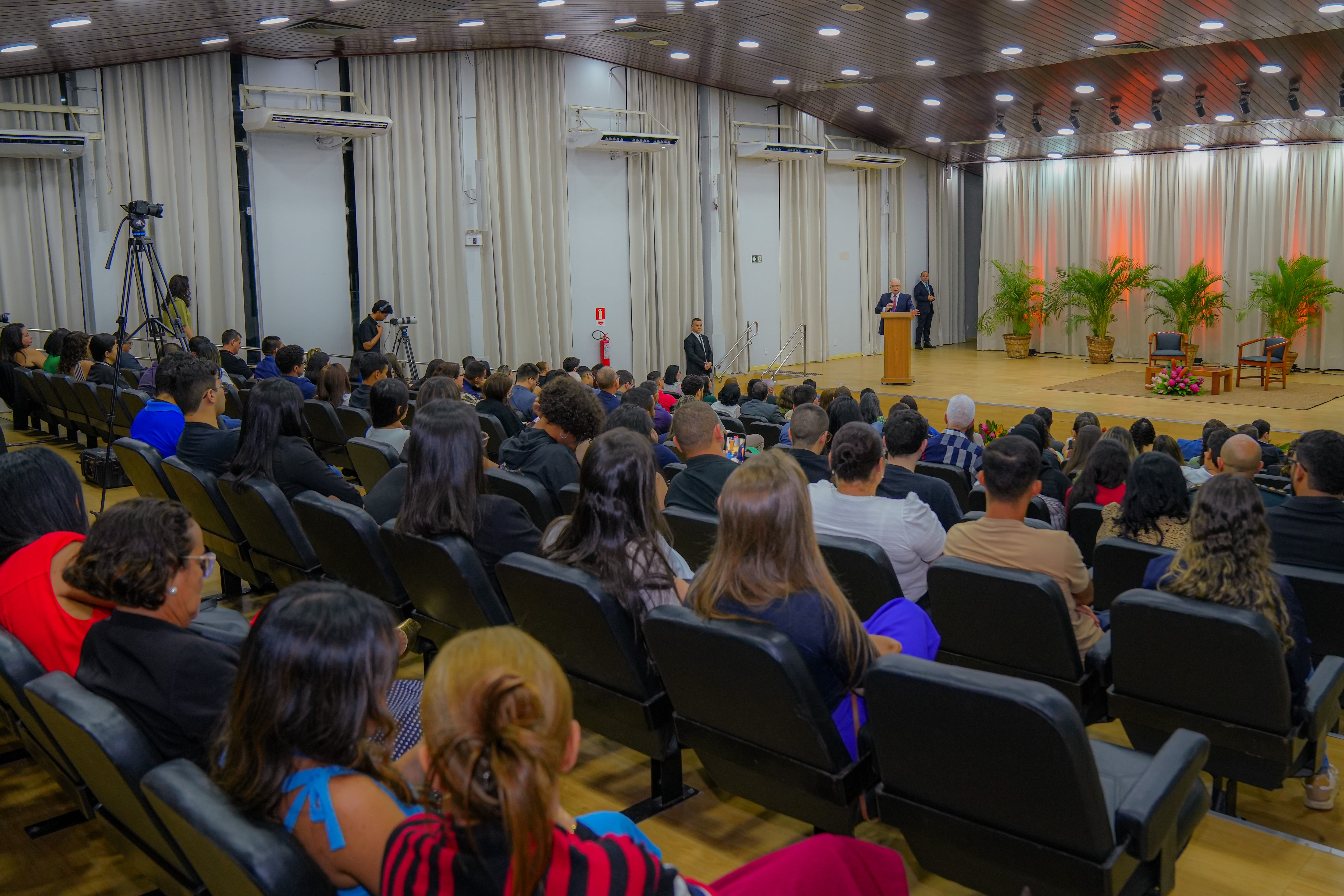 A imagem mostra o Ministro Edson Fachin discursando em um auditório, em pé atrás de um púlpito no centro do palco. A plateia, vista de costas, assiste atenta. O ambiente é formal, com cortinas claras e iluminação suave.