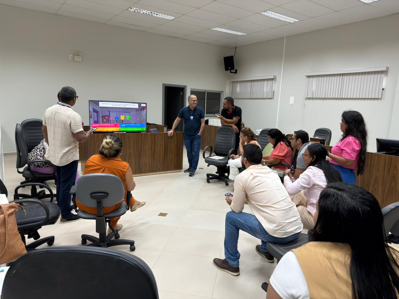 A imagem mostra uma sessão de treinamento. Um homem de camisa azul e óculos conduz a fala, enquanto outro aponta para uma TV que exibe gráficos. Participantes, sentados em cadeiras de escritório, acompanham a atividade. O ambiente é claro, organizado e profissional. A imagem mostra uma sessão de treinamento. Um homem de camisa azul e óculos conduz a fala, enquanto outro aponta para uma TV que exibe gráficos. Participantes, sentados em cadeiras de escritório, acompanham a atividade. O ambiente é claro, organizado e profissional.