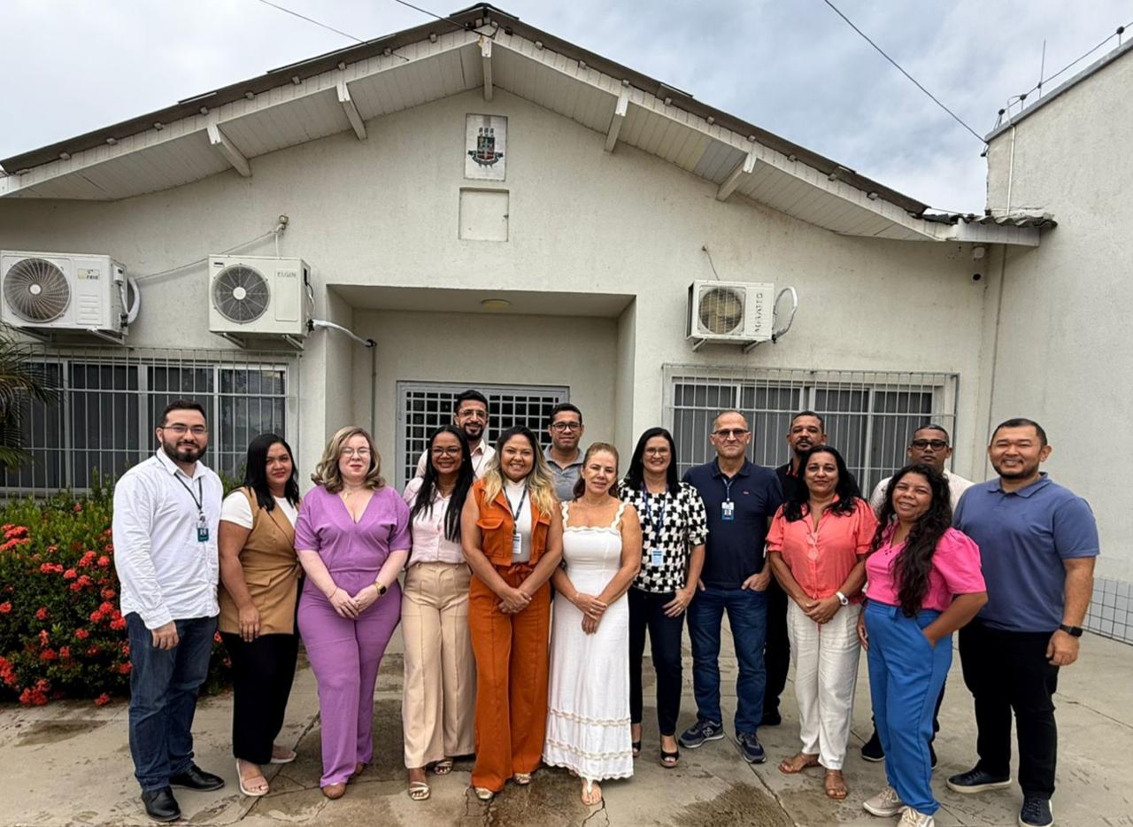 A imagem mostra um grupo de 14 pessoas posando para foto em frente a um edifício tradicional de cor clara, com telhado triangular e brasão na fachada. Vestidos de forma casual a semi-formal, os participantes demonstram clima descontraído, indicando um encontro ou reunião. O cenário inclui plantas floridas e céu nublado. A imagem mostra um grupo de 14 pessoas posando para foto em frente a um edifício tradicional de cor clara, com telhado triangular e brasão na fachada. Vestidos de forma casual a semi-formal, os participantes demonstram clima descontraído, indicando um encontro ou reunião. O cenário inclui plantas floridas e céu nublado.