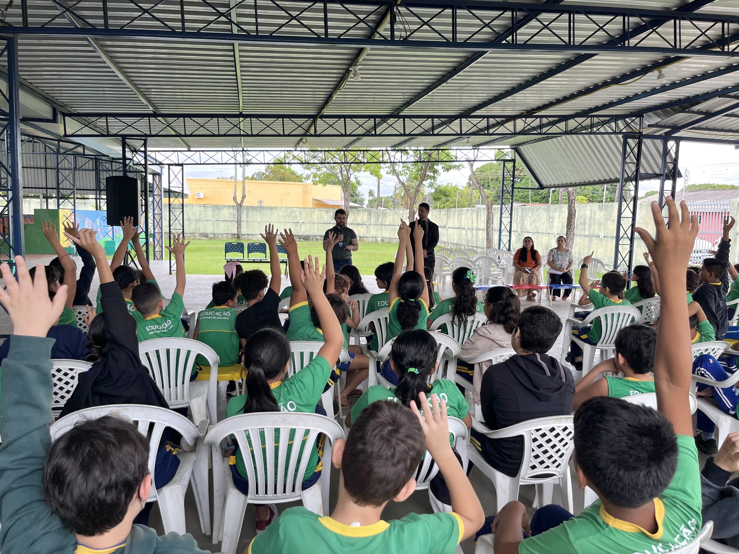 A imagem retrata um evento educativo em um espaço coberto e aberto, com várias crianças usando camisetas verdes, sentadas em cadeiras plásticas brancas. Muitas levantam os braços, participando ativamente. Ao fundo, adultos conduzem a atividade, com um deles usando microfone. O ambiente é simples e cercado por natureza. A imagem retrata um evento educativo em um espaço coberto e aberto, com várias crianças usando camisetas verdes, sentadas em cadeiras plásticas brancas. Muitas levantam os braços, participando ativamente. Ao fundo, adultos conduzem a atividade, com um deles usando microfone. O ambiente é simples e cercado por natureza.