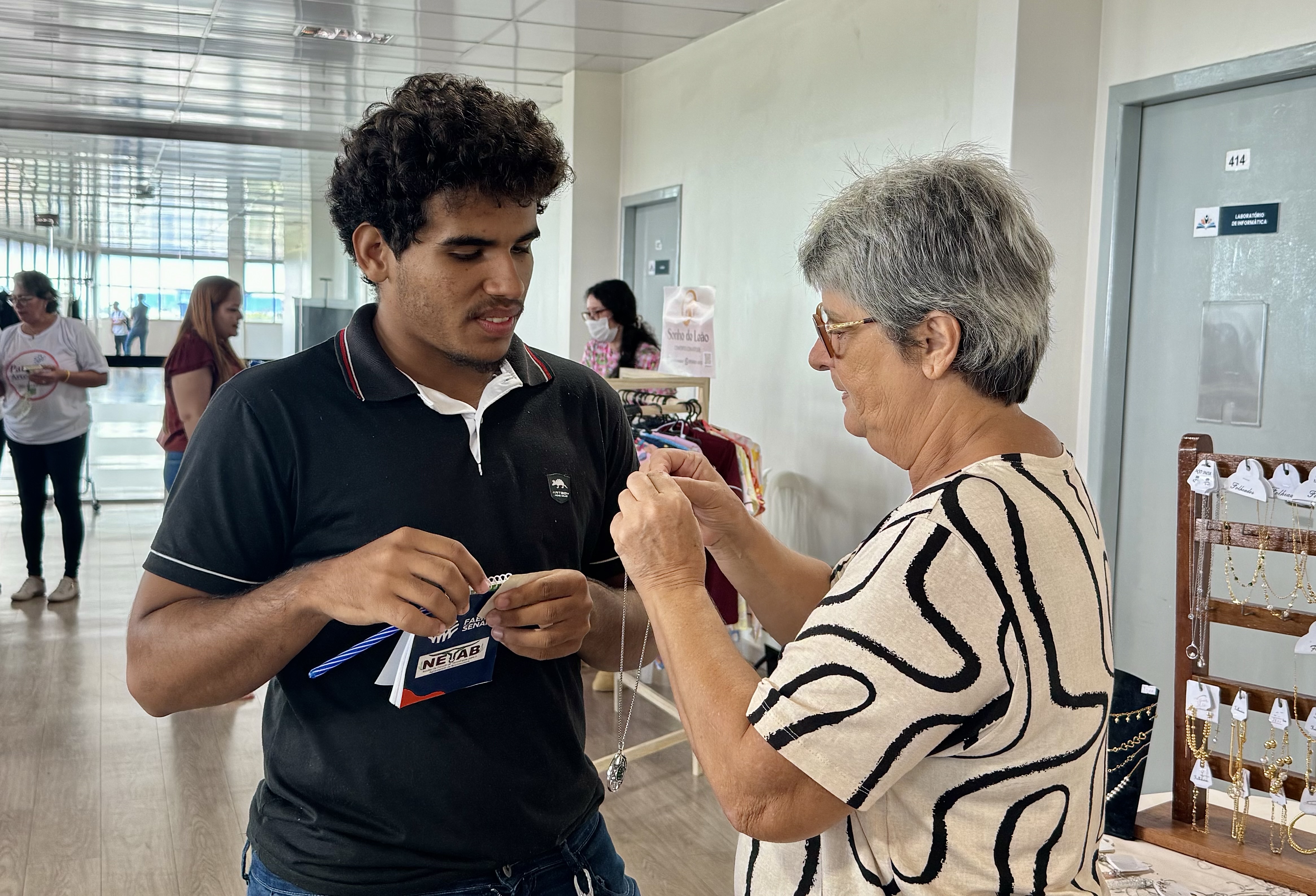 Imagem colorida mostra uma das empreendedoras interagindo com um cliente em frente a uma mesa de exposição com colares e pulseiras. Imagem colorida mostra uma das empreendedoras interagindo com um cliente em frente a uma mesa de exposição com colares e pulseiras.