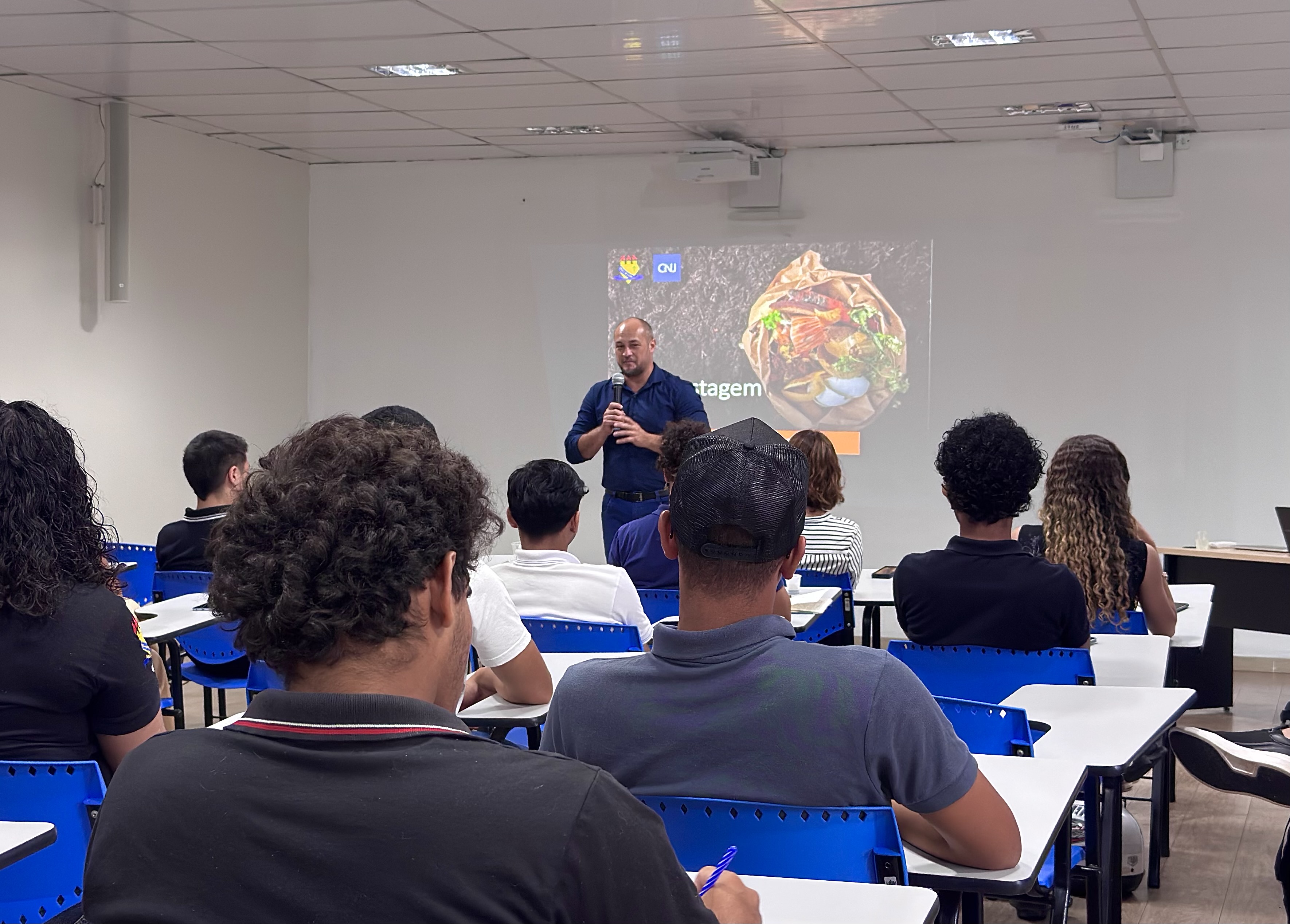 Imagem colorida mostra uma sala de aula com pessoas sentadas em carteiras, assistindo à apresentação do palestrante Diego Cruz. No fundo, um slide projetado na parede exibe imagens relacionadas à compostagem. Imagem colorida mostra uma sala de aula com pessoas sentadas em carteiras, assistindo à apresentação do palestrante Diego Cruz. No fundo, um slide projetado na parede exibe imagens relacionadas à compostagem.