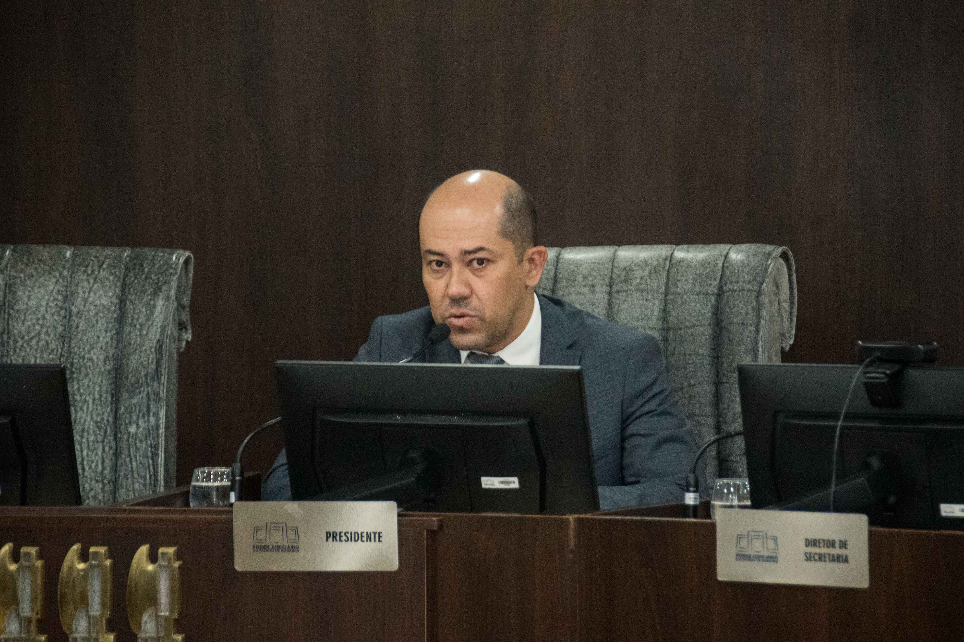 Homem calvo, de terno escuro, sentado em uma cadeira de presidente em um plenário. Ele fala ao microfone e, sobre a mesa de madeira à sua frente, há uma placa de identificação com a palavra "PRESIDENTE". Homem calvo, de terno escuro, sentado em uma cadeira de presidente em um plenário. Ele fala ao microfone e, sobre a mesa de madeira à sua frente, há uma placa de identificação com a palavra "PRESIDENTE".