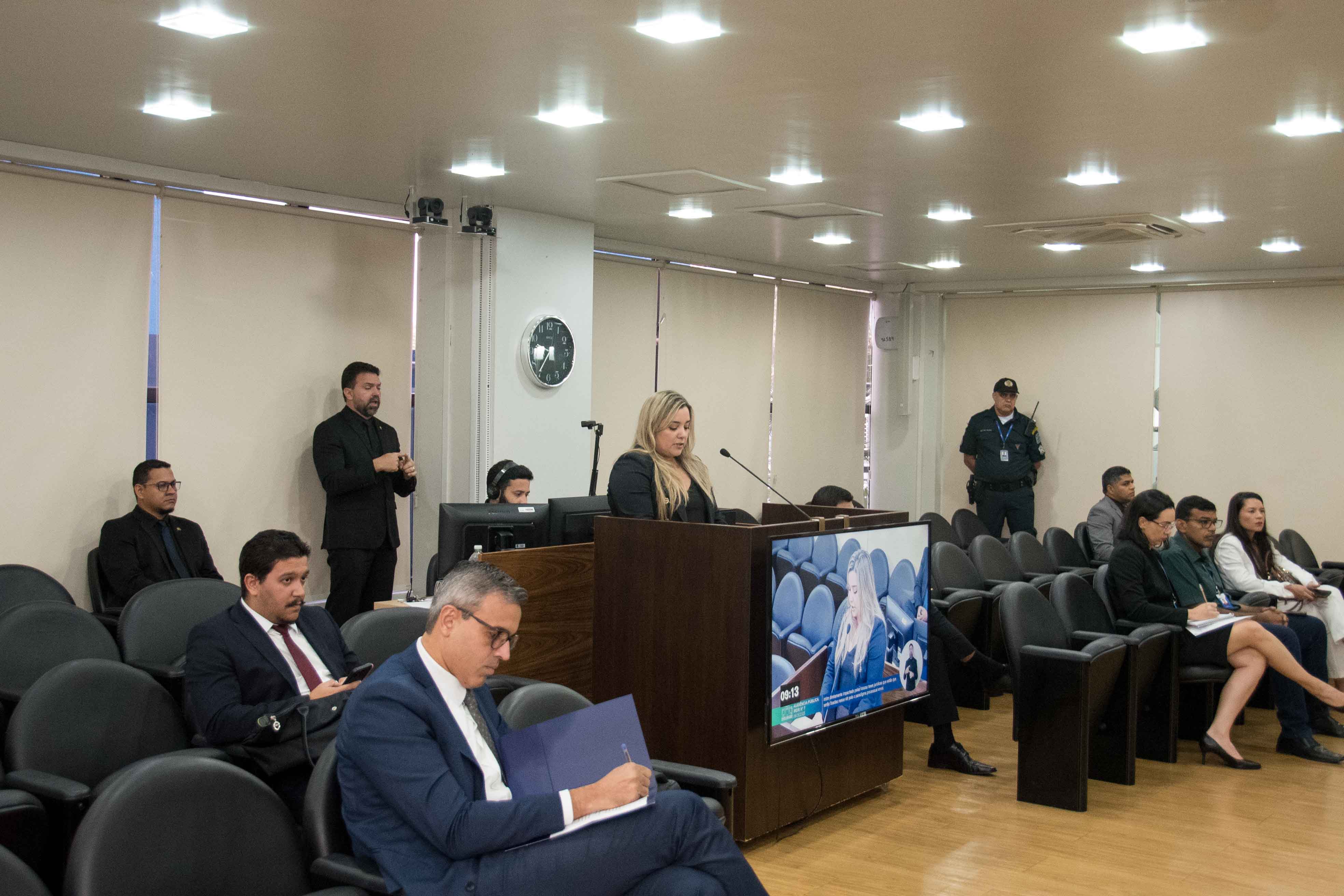 Foto de um auditório ou sala de plenário. Ao centro, uma mulher loira está de pé em uma tribuna de madeira, discursando. À sua frente, uma plateia de homens e mulheres com roupas formais assiste, sentada em cadeiras escuras. Em primeiro plano, um homem de óculos e terno anota em uma pasta.