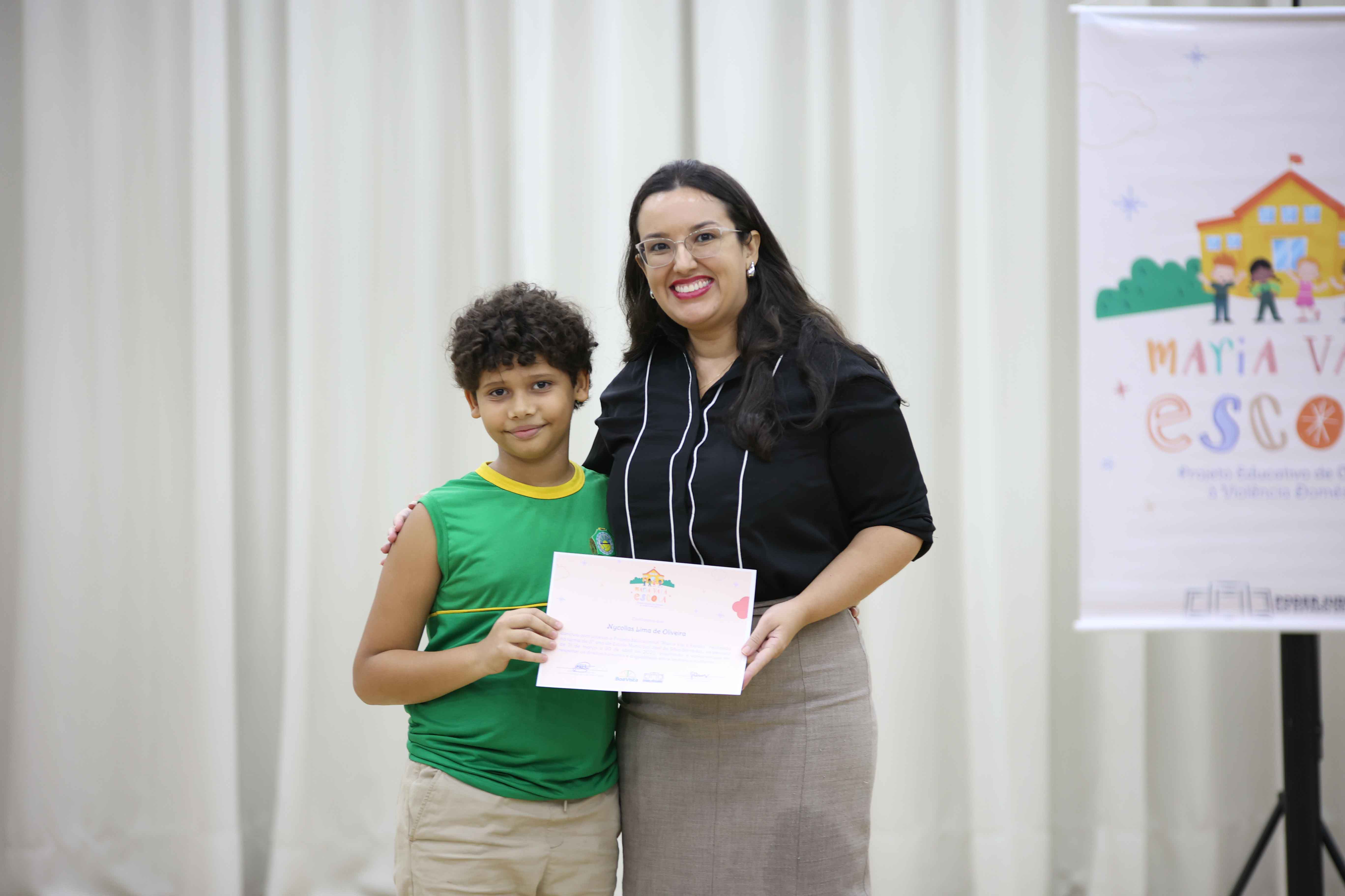 Foto colorida contém a juíza Suelen Alves, coordenadora da Coordenadoria Estadual da Mulher em Situação de Violência Doméstica (Cevid/TJRR), posando para fotografia ao lado de 15 crianças e mais quatro mulheres,  durante a certificação do Maria Vai à Escola. 