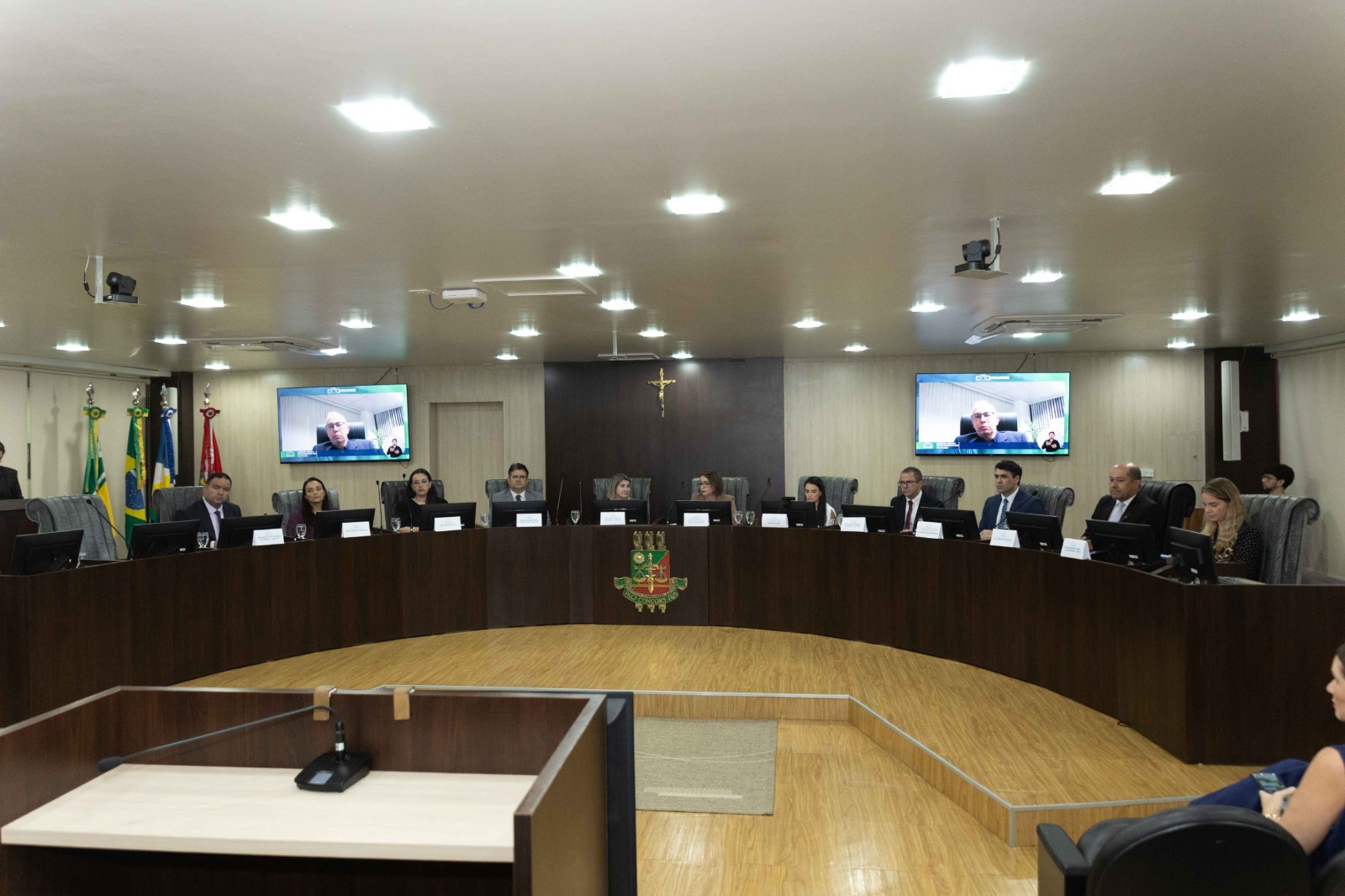 Pessoas sentadas em uma mesa em formato de “U” dentro de um plenário, com computadores à frente. No centro da parede há um crucifixo e o brasão do TJRR. Duas telas exibem participantes por videoconferência. Pessoas sentadas em uma mesa em formato de “U” dentro de um plenário, com computadores à frente. No centro da parede há um crucifixo e o brasão do TJRR. Duas telas exibem participantes por videoconferência.